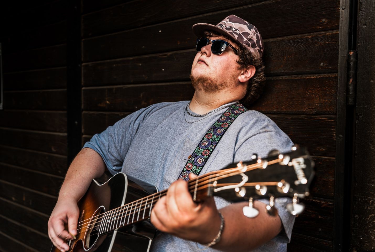 A man leaning against a wooden wall holding a guitar wearing a blue tshirt, camo hat, and black sunglasses